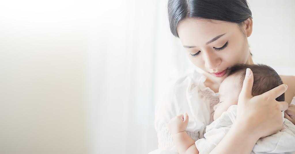 Maternal Mental Health Awareness Month - Asian woman cradling a baby in her arms, gazing down at the child with love.