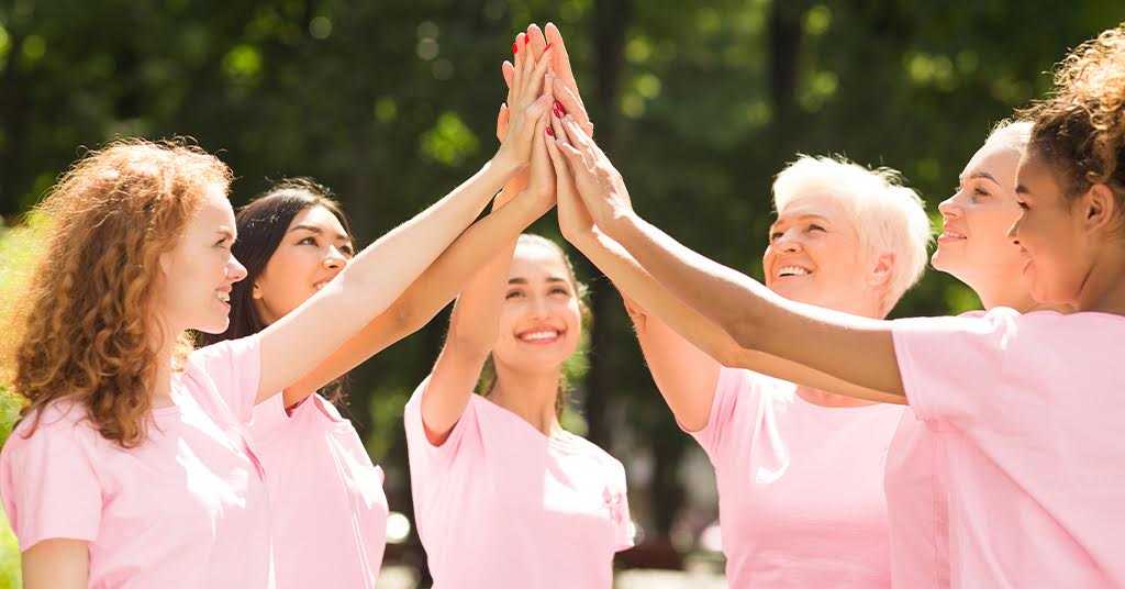 Group of multiethnic women wearing pink t-shirts giving high fives outdoors representing cancer awareness and mental health support.