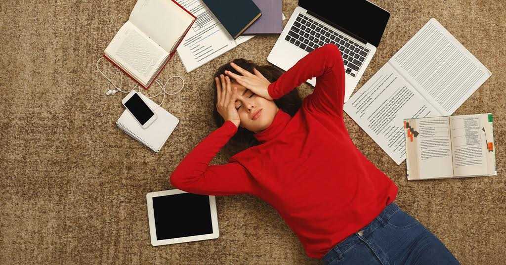 Teen Exam Anxiety: adolescent student wearing a red jumper lying on the floor amidst scattered books and digital devices.