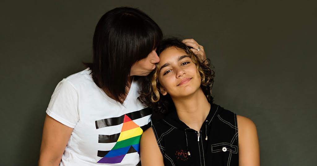 LGBTQ teen support - mother wearing a white t-shirt with a LGBTQ+ flag hugging teenager.