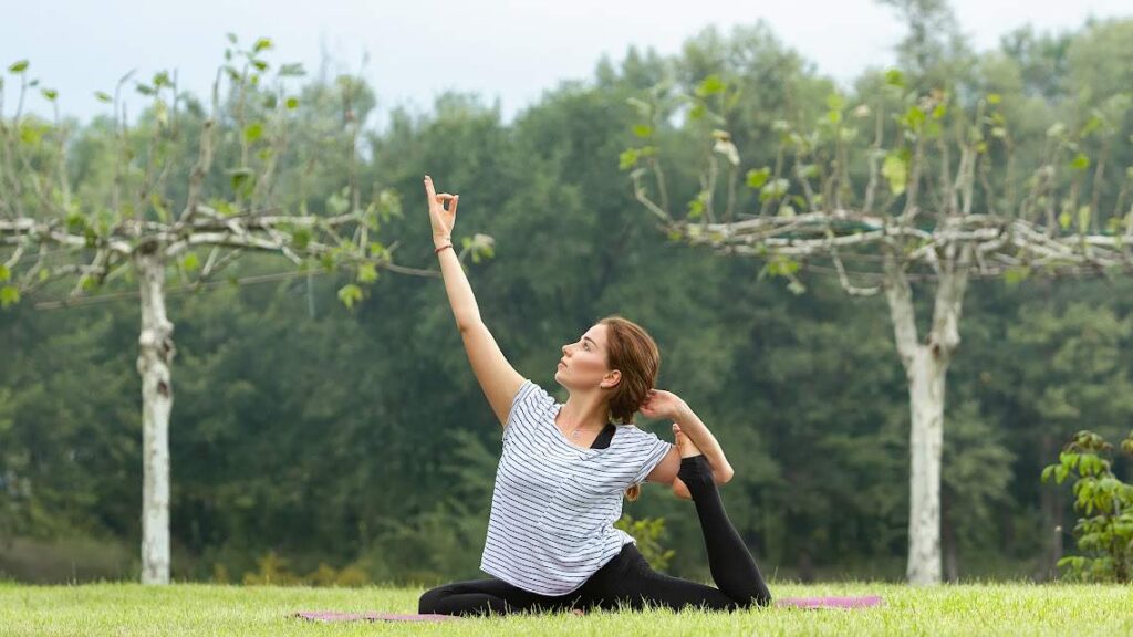 Photo of a woman in a yoga pose, gracefully balancing on one leg in a lush garden. She’s surrounded by vibrant greenery and radiating calm.