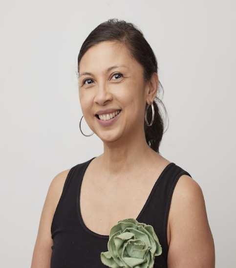 A smiling woman with black hair, dressed in a black dress, adorned with a green bow. She exudes elegance and cheerfulness against a neutral background.