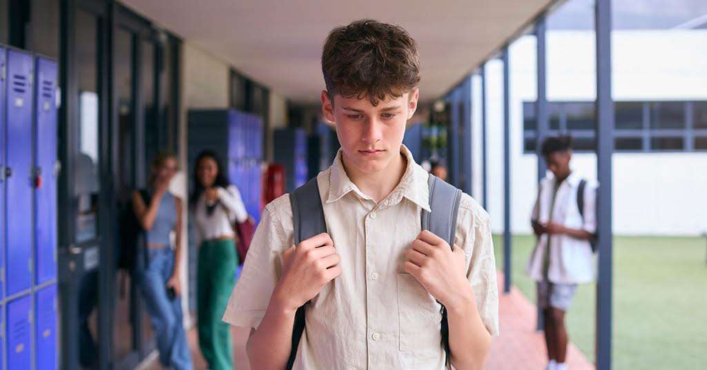 A college student with a sad expression walks away from a group of peers who are teasing him, highlighting the emotional impact of bullying and social exclusion.