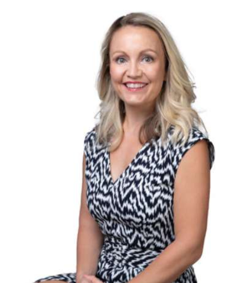 Smiling lady with blonde hair, dressed in a stylish black and white outfit. She exudes confidence and warmth, set against a simple, neutral background.