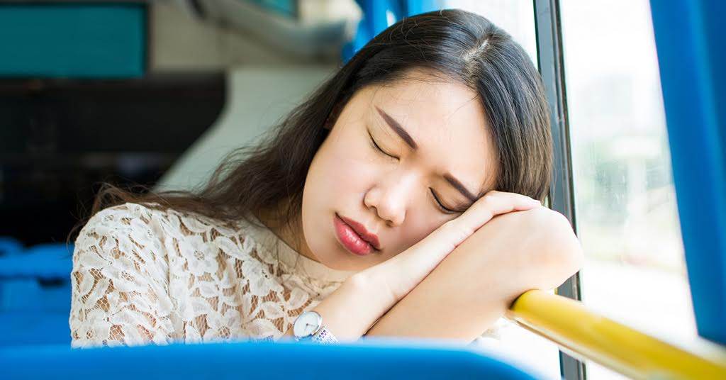 An Asian woman asleep on a bus. She's leaning against the window, her head resting on her folded arms. It is a quiet moment of rest experienced on a public bus.