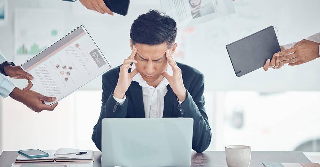 A man sitting at his desk, visibly stressed and exhausted, with his hands on his head, overwhelmed by work pressure