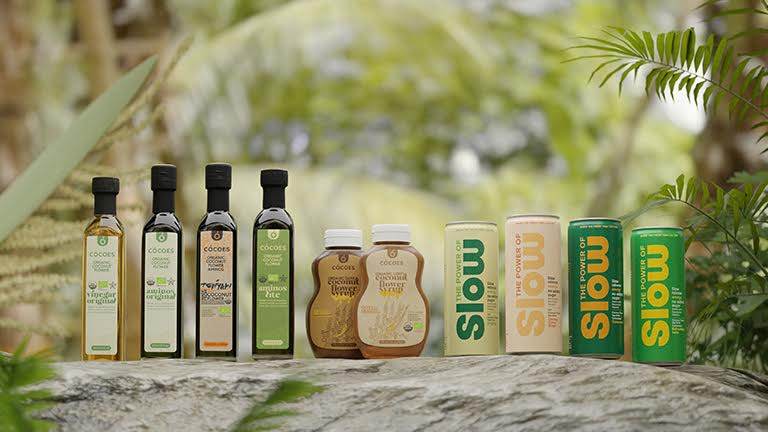 A variety of CÓCOES products are displayed on a wooden surface in a lush green setting. The products include organic oils, maple syrup, and energy drinks.