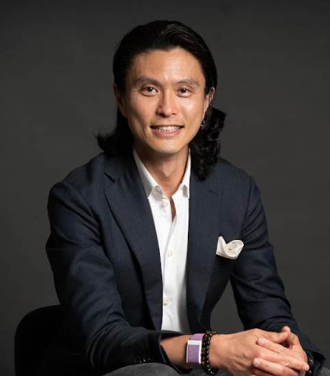 A man in a blazer and a white collared shirt sits against a dark grey background. The man's body is angled slightly toward the camera, and his posture is relaxed but attentive.