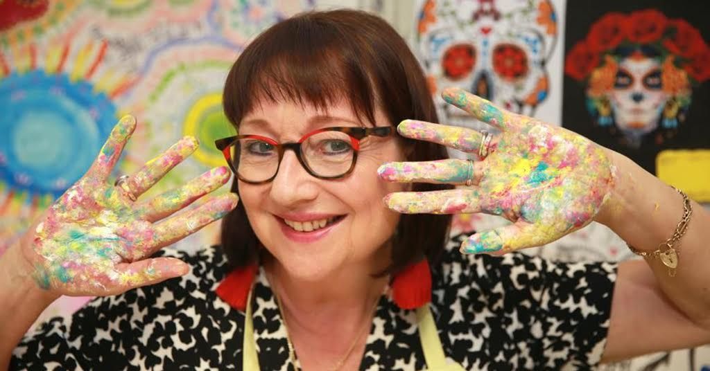 A cheerful woman wearing glasses and a yellow apron. Her hands are raised and covered in colorful paint, she is engaging in artistic activities.