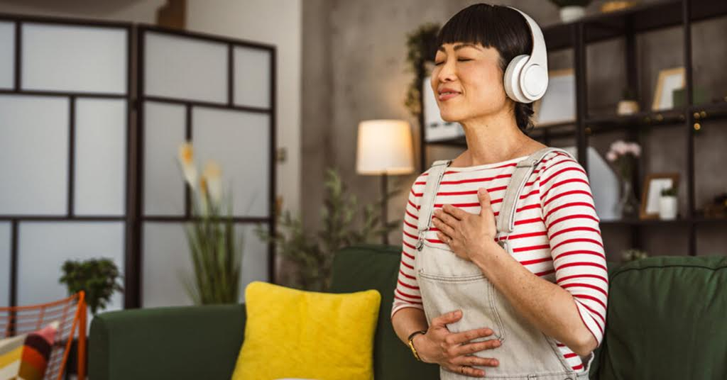 A woman wearing headphones with and listening to music calmly with her eyes closed and hands relaxing calmly on her chest