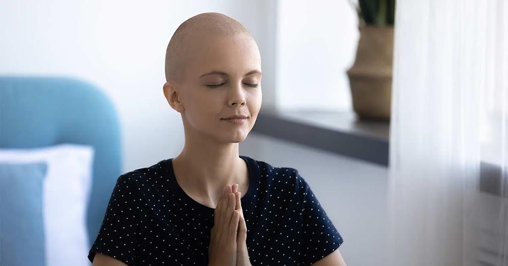 A serene woman, likely a cancer patient, meditates with closed eyes and hands in prayer. The bright room and soft light evoke peace and mindfulness.