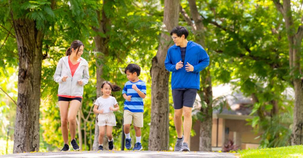 An image of a family of 4 jogging happily in a park, their faces lit up with smiles and energy. The parents are encouraging their kids, who are full of excitement and laughter.