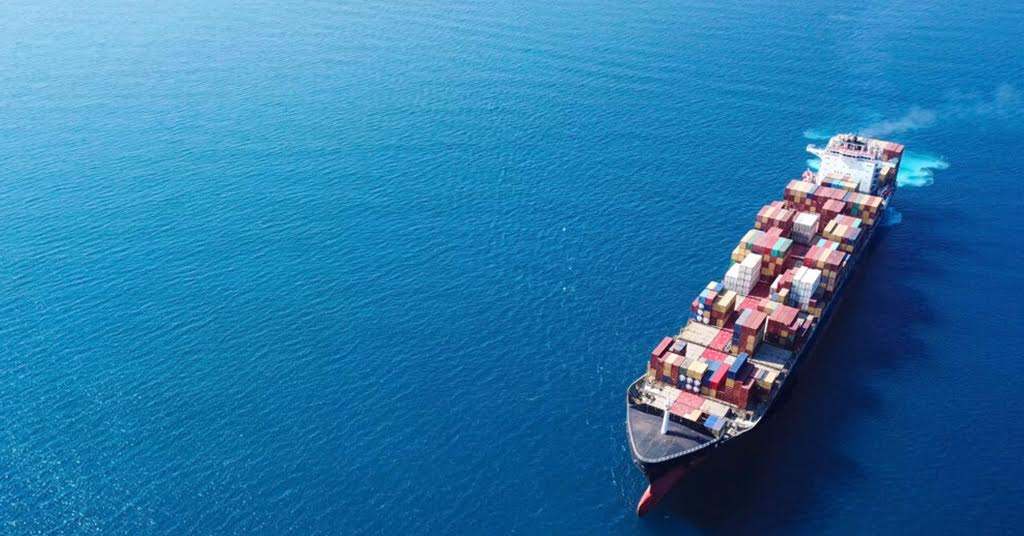 The image shows a large cargo ship sailing on a vast ocean. The ship is loaded with numerous colorful shipping containers stacked high on its deck.